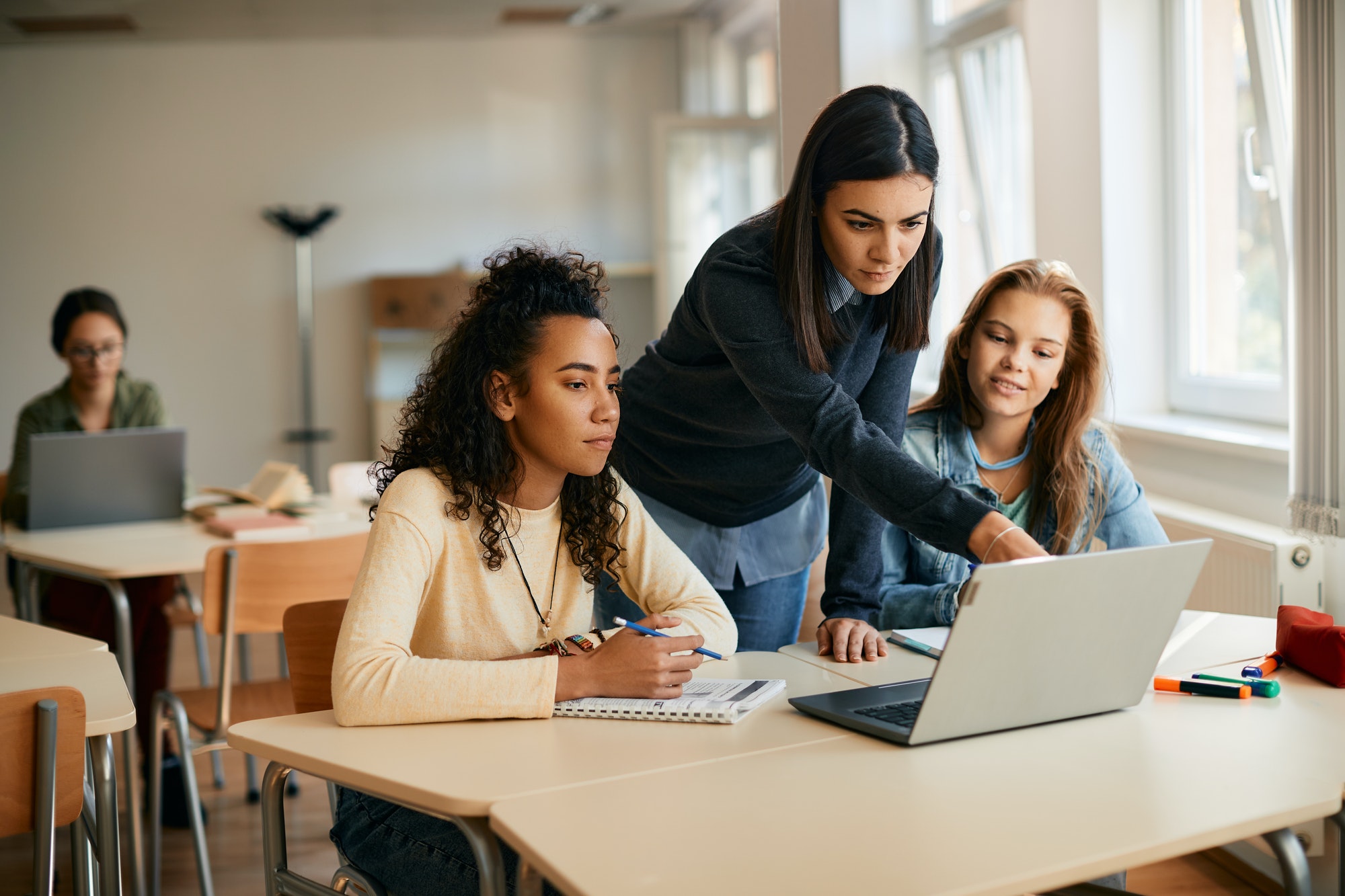 High school professor assisting her students in e-learning on laptop in the classroom.